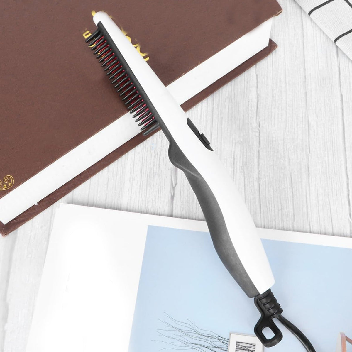 A white and black electric hair and beard straightening comb rests on an open book and a patterned sheet.