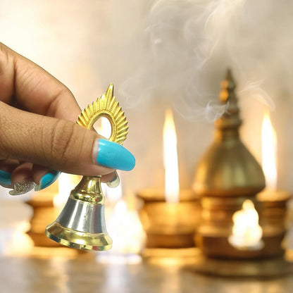 A hand with blue nail polish holds the small brass and steel pooja bell, with blurred lit diyas and smoke in the background,