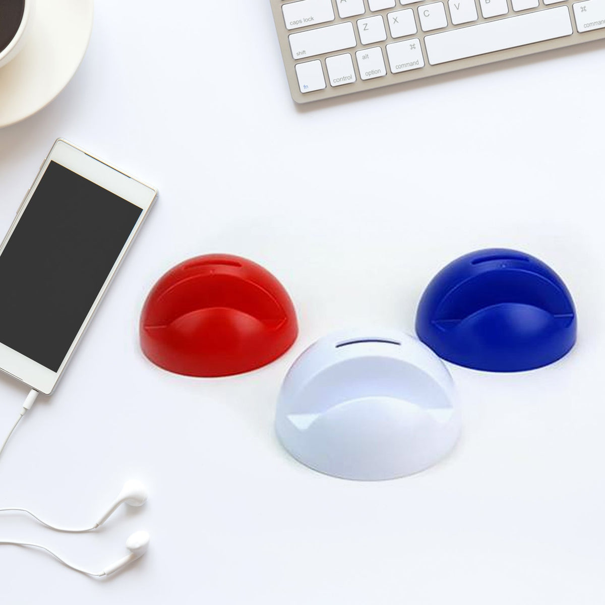 A white phone with earbuds, and red, white, and blue phone holder piggy banks on a white desk.