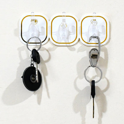 Three transparent adhesive hooks with gold borders on a white wall, holding two sets of keys.