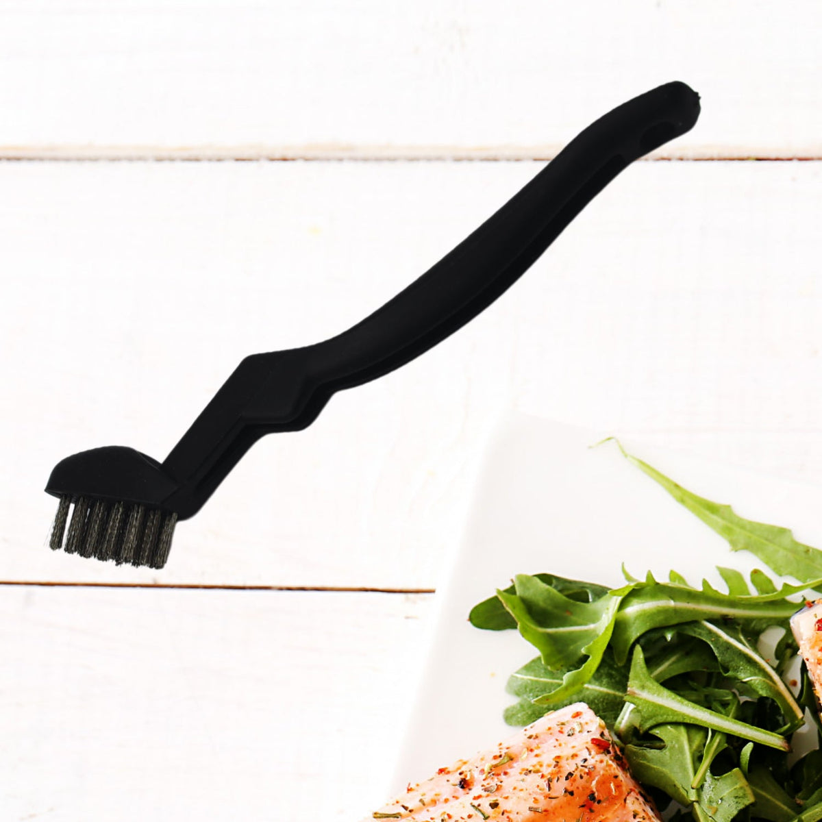 A black grill cleaning brush with metal bristles rests on a white wooden surface next to a plate of food.