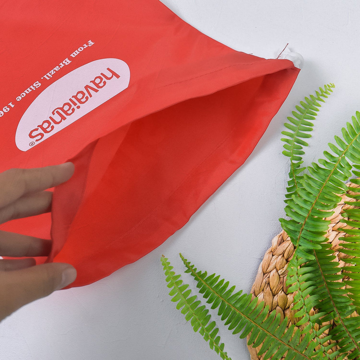 A hand opening a red drawstring bag with the Havaianas logo, next to a woven mat and green fern leaves.