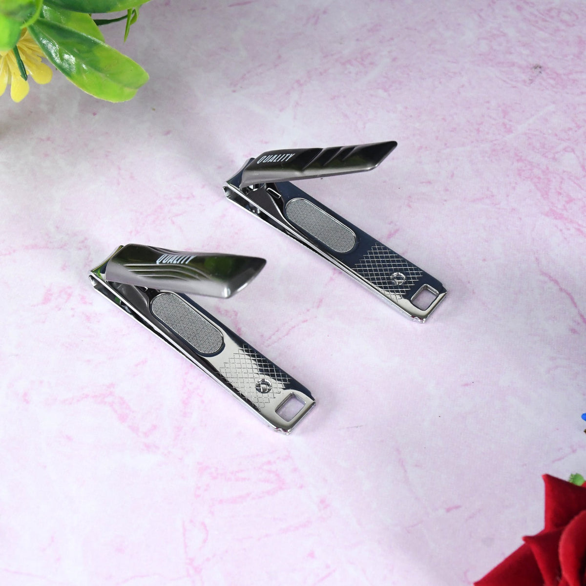 Two black and silver nail clippers, partially open, on a pink marble surface with a red flower in the corner.