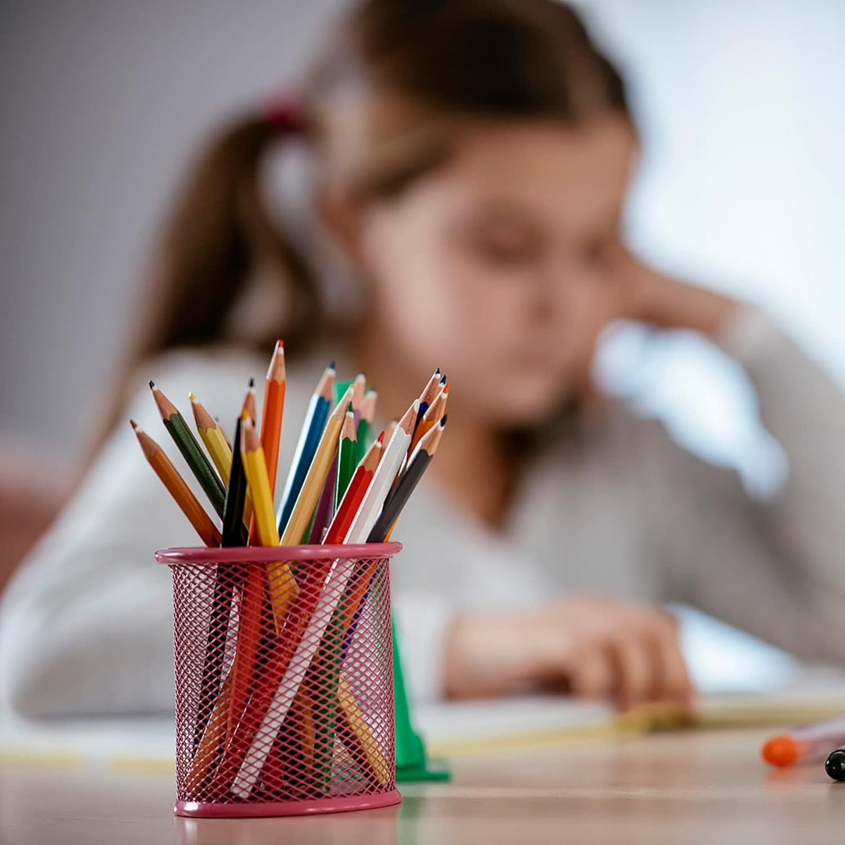 A pink metal mesh pen holder filled with pencils sits on a desk with a blurred background of a girl drawing.