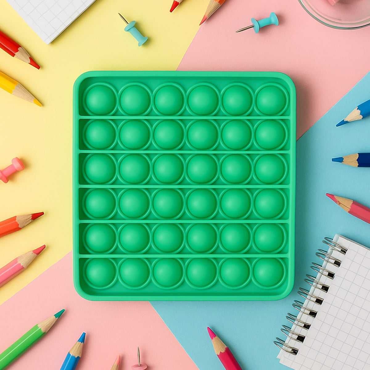 A square green silicone pop-it fidget toy surrounded by colorful pencils and thumbtacks on a multi-colored background.