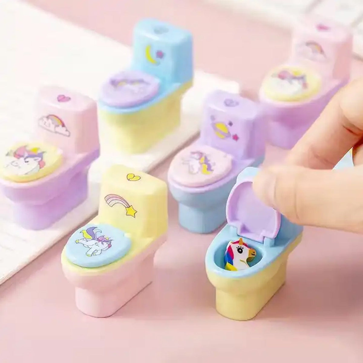 A hand opens a pastel blue and yellow toilet-shaped sharpener to reveal a small unicorn eraser inside, surrounded by other