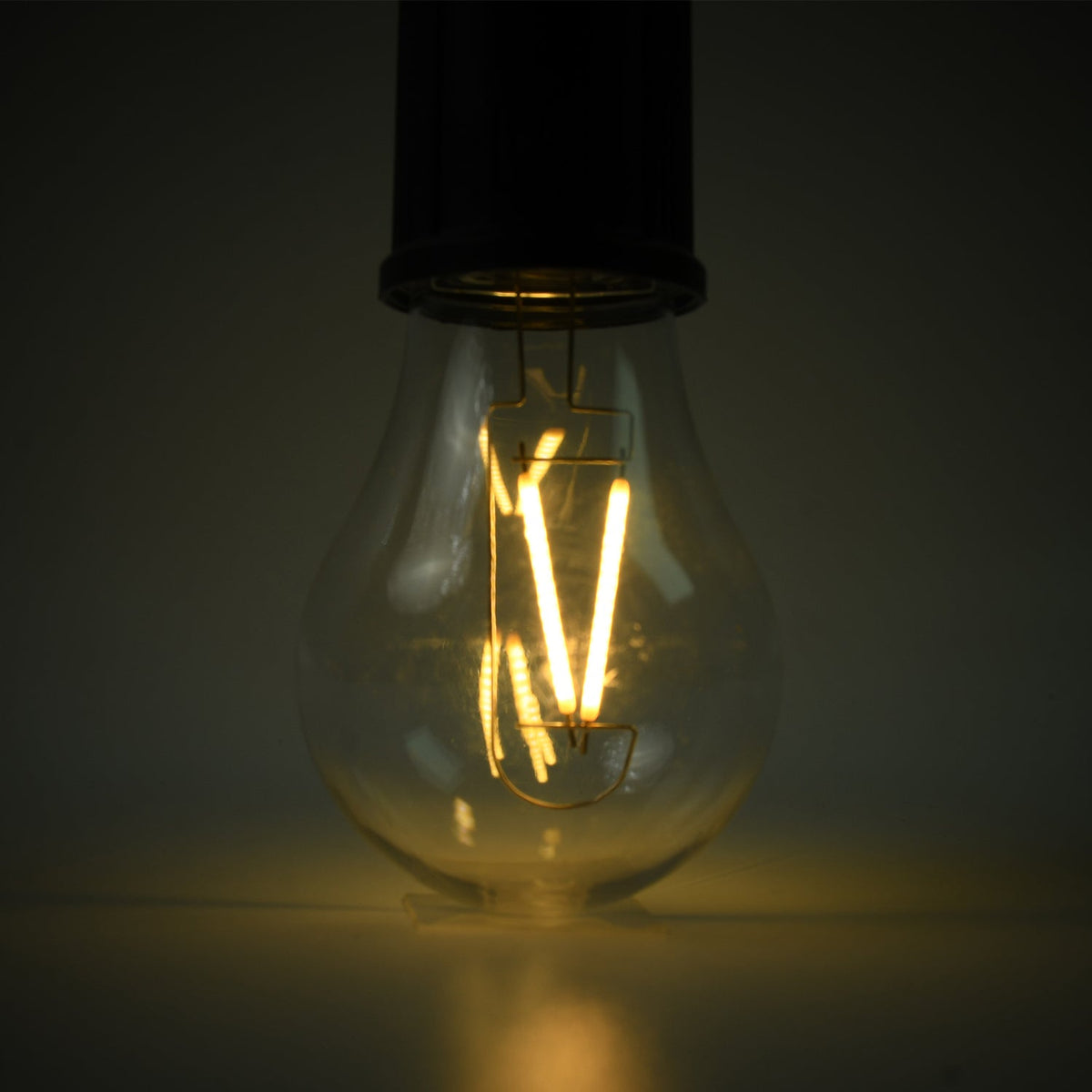 A close-up of the clear LED bulb shows its warm yellow filaments glowing brightly against a dark background.