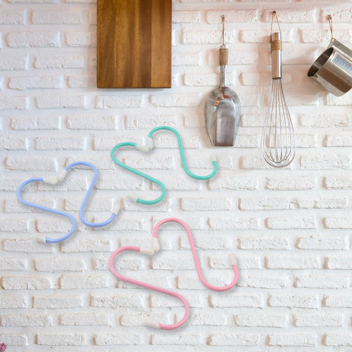Three colorful plastic coated S hooks, blue, green, and pink, hanging on a white brick wall.