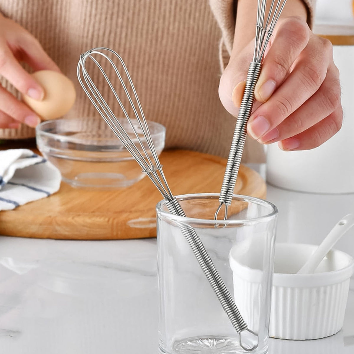 Two small silver whisks stand upright in a clear glass, with a person and an egg in the blurred background.
