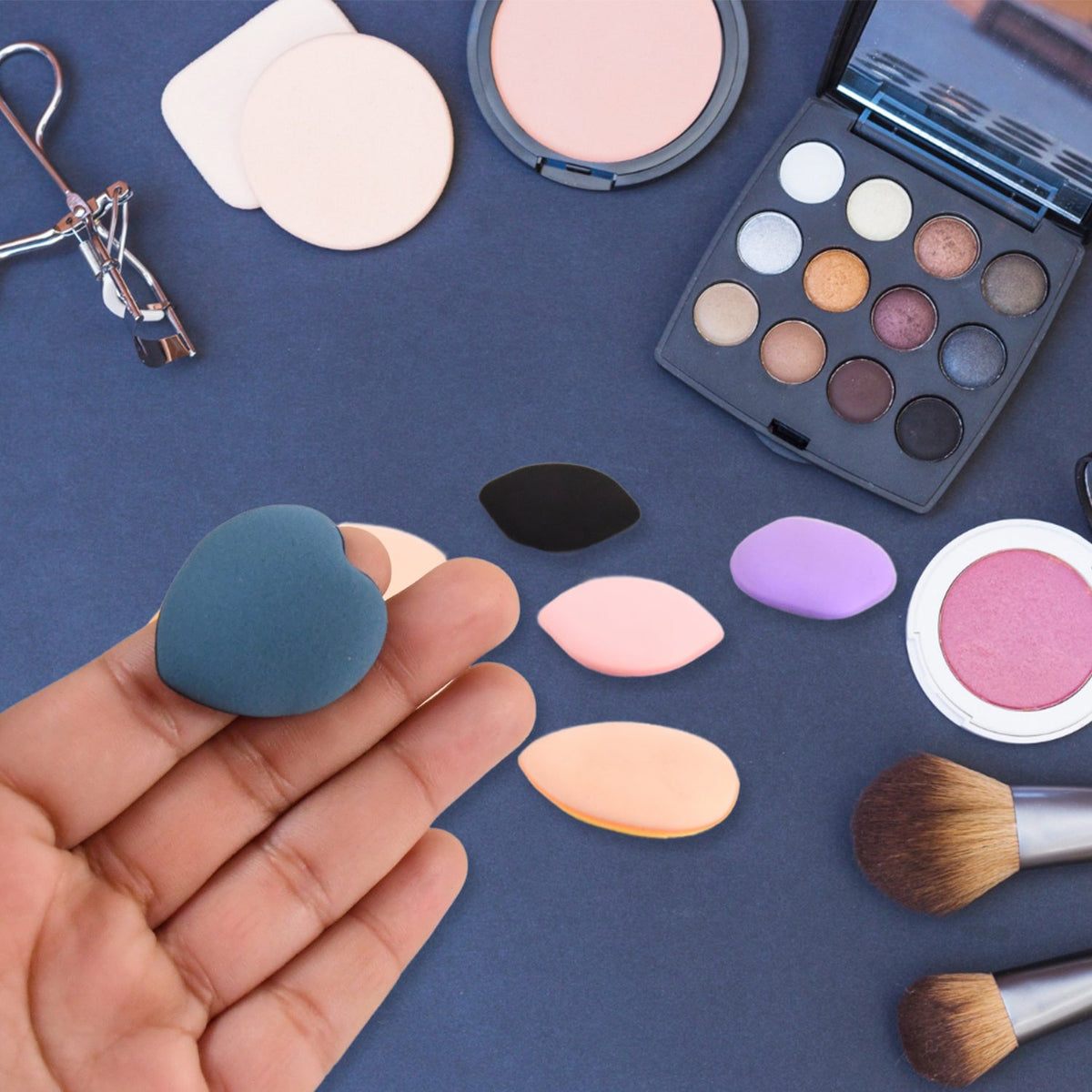 A hand holding a blue heart-shaped makeup sponge, surrounded by other sponges and makeup tools on  blue surface