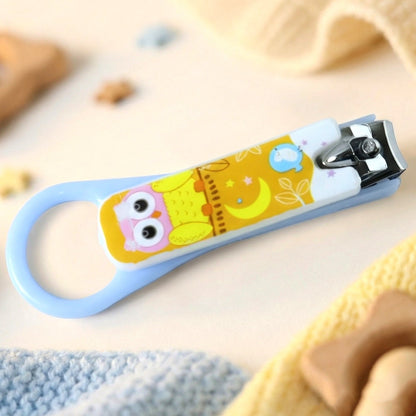 A light blue baby nail clipper with a yellow owl design, resting on a soft, light-colored blanket with toys nearby.