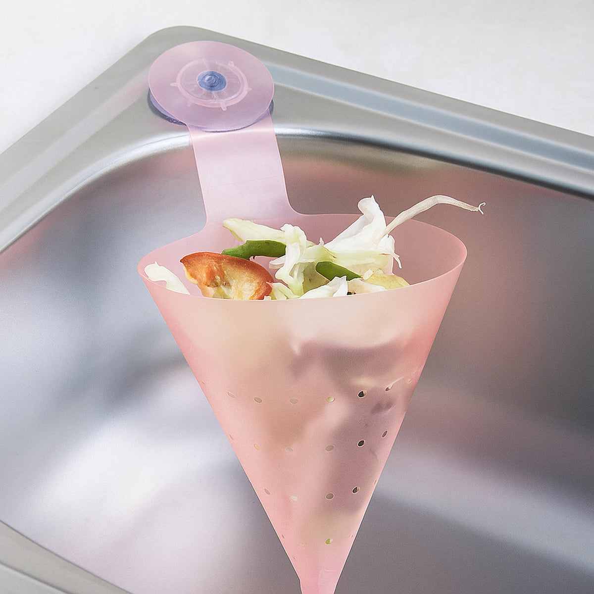 A pink silicone sink strainer with a suction cup, holding vegetable scraps, attached to a metal sink.