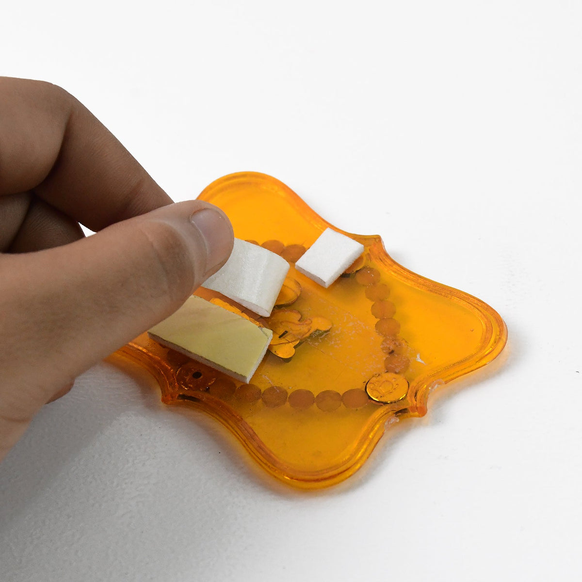 A hand applying white adhesive foam squares to the back of a yellow Shubh Labh wall hanging.