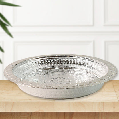 A silver-colored plastic pooja thali with embossed patterns, sitting on a light wooden surface with a white paneled wall
