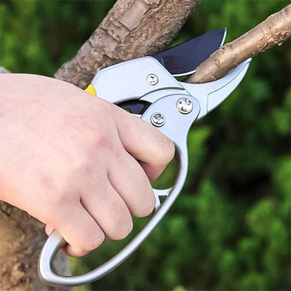 A hand holding silver and yellow pruning shears, cutting a small tree branch.