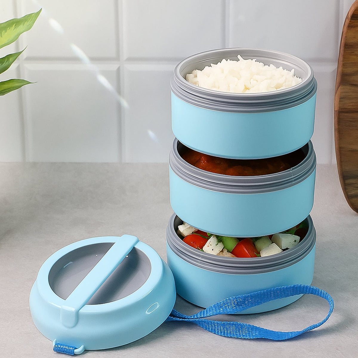 The light blue three-layer lunch box is open, revealing rice, curry, and salad in its compartments, with the lid beside it.