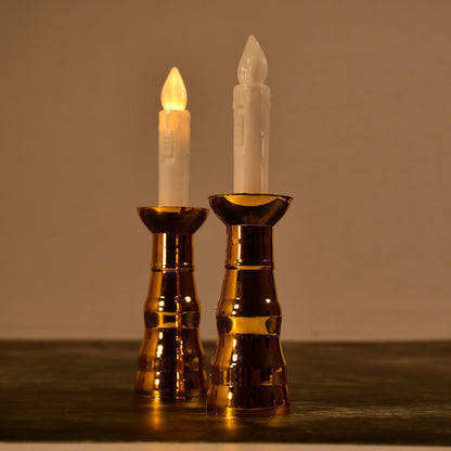 Two gold-based LED candles with illuminated flames stand on a dark surface in a dimly lit room.