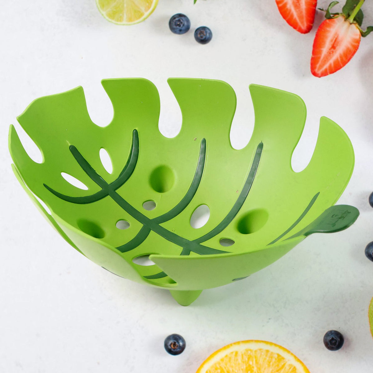 A green leaf-shaped plastic fruit basket on a white surface surrounded by various fruits like blueberries and strawberries.