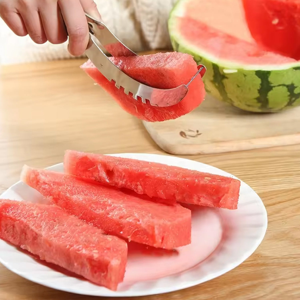 A hand uses a stainless steel watermelon slicer to serve a piece of watermelon onto a white plate with other slices.