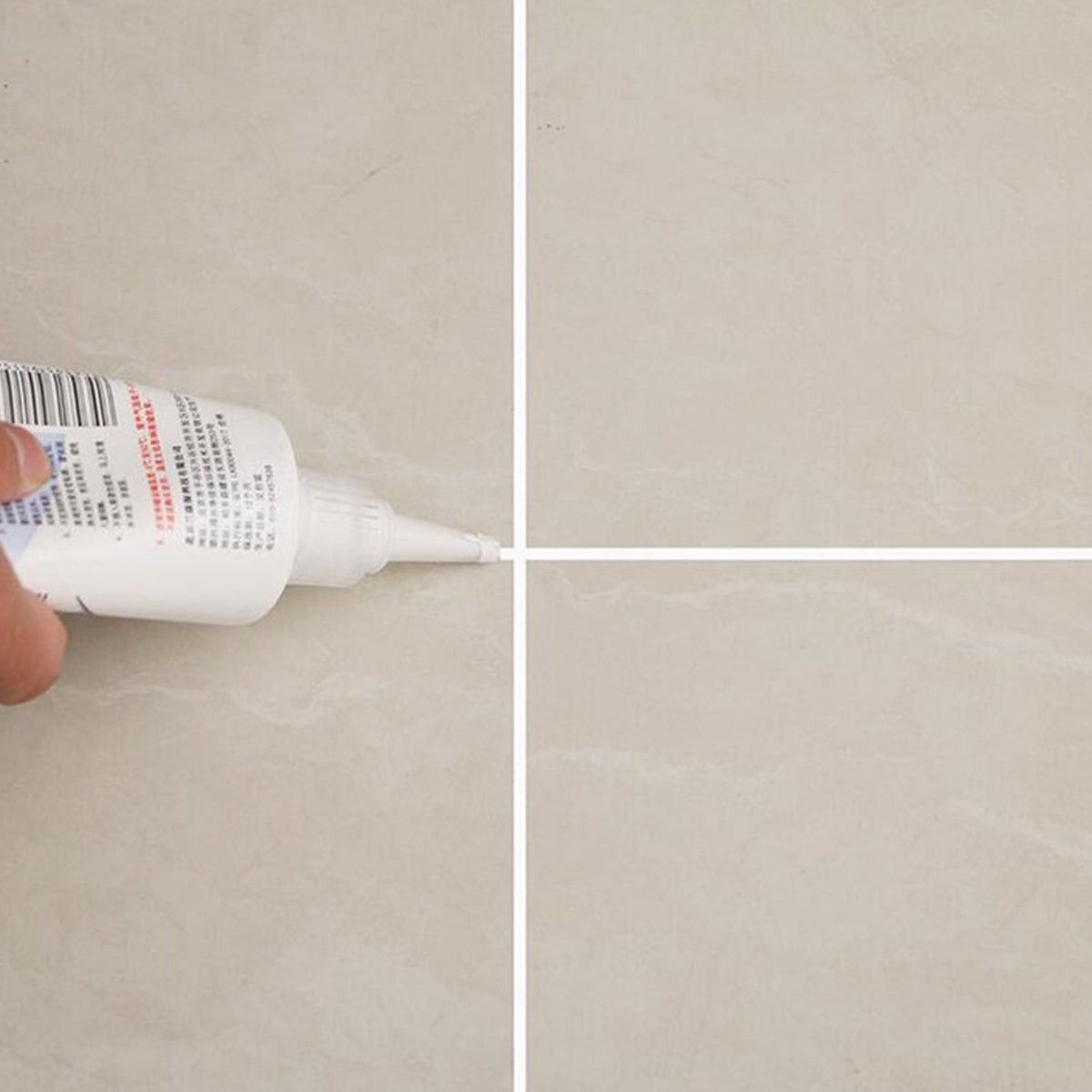 Close-up of a hand applying white sealant from a tube to the intersection of four beige floor tiles.