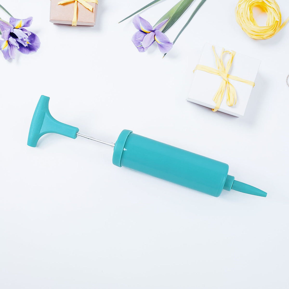 A teal manual air pump with a silver rod, surrounded by purple flowers, yellow string, and small gift boxes on a white
