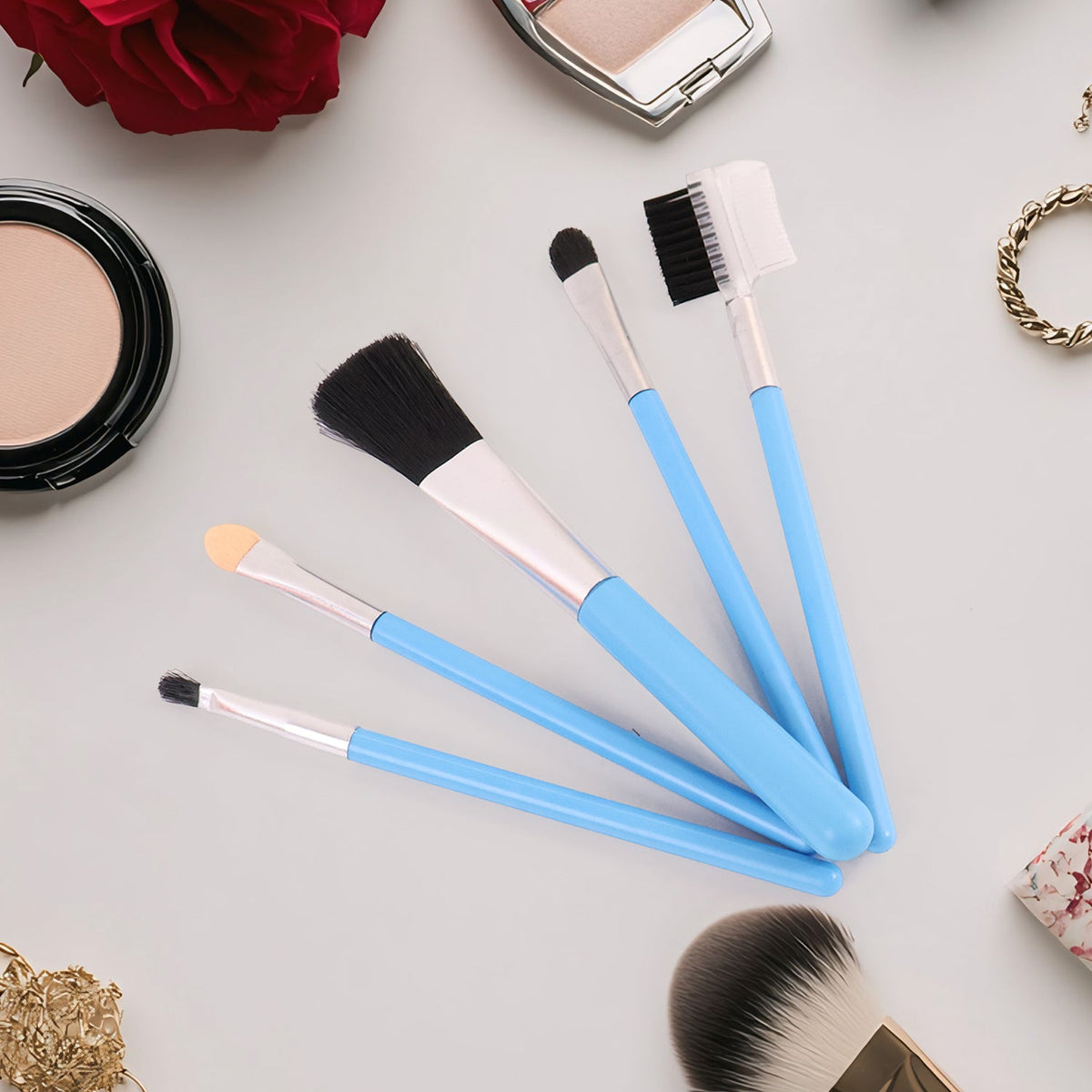 Five light blue makeup brushes are fanned out on a light grey surface, surrounded by various makeup items.
