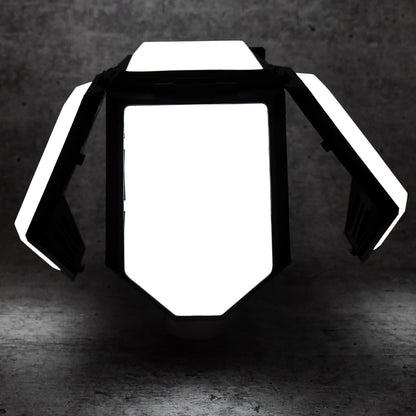 The white and black foldable LED lantern with its panels partially open, glowing brightly in a dark setting.