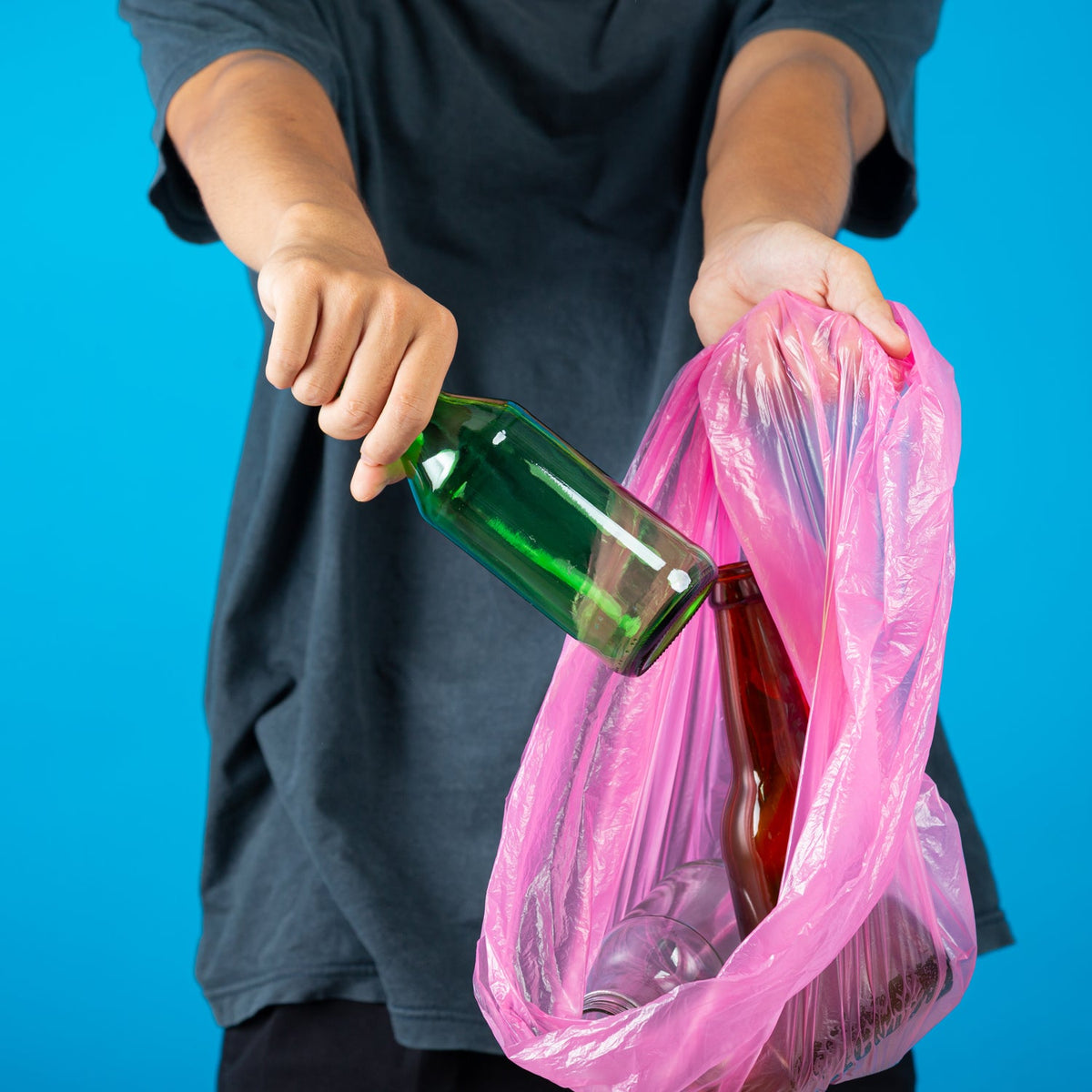 A person in a dark shirt puts a green glass bottle into a pink plastic garbage bag.