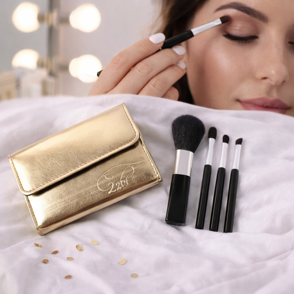 A gold makeup brush pouch and four black and silver brushes on a white sheet, with a woman applying eyeshadow in the