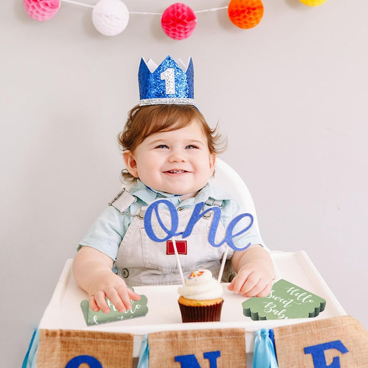 A smiling baby in a high chair, wearing a blue crown, with mint green scalloped napkins beside a cupcake.