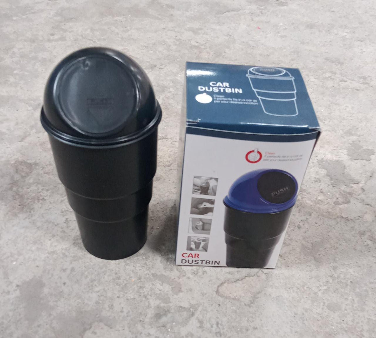 A black car dustbin with a black lid stands next to its product box on a concrete surface.