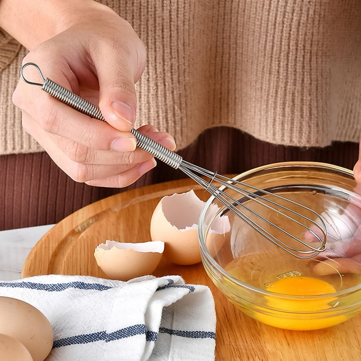A person in a beige sweater holds a small silver whisk over a clear glass bowl with an eggshell nearby.