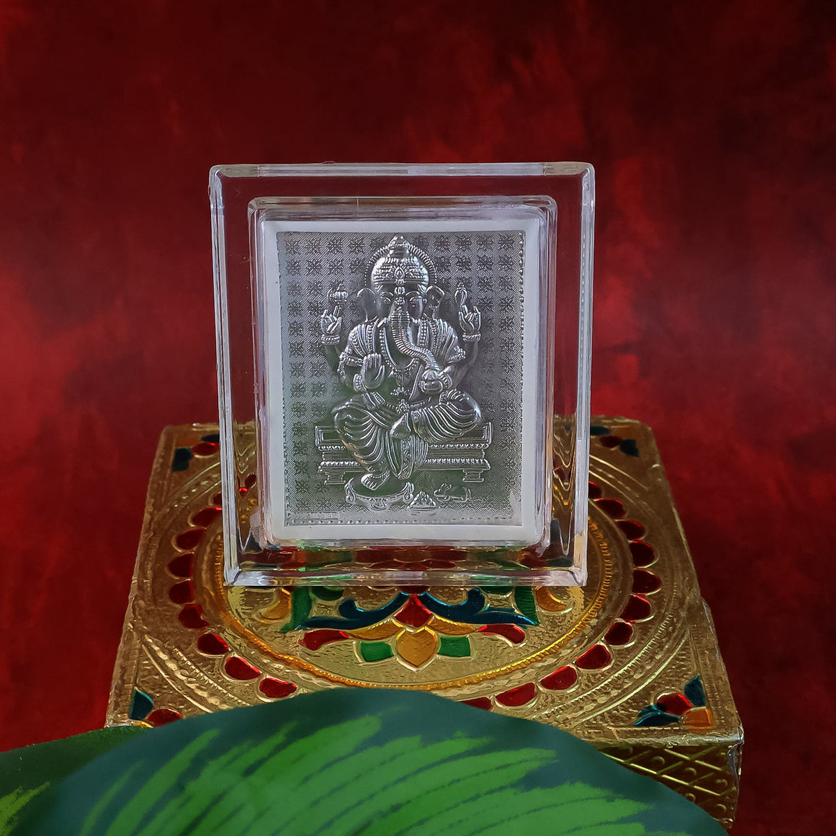 Silver-plated Ganesha idol in an acrylic frame, placed on an ornate gold and multicolored box with a green leaf.