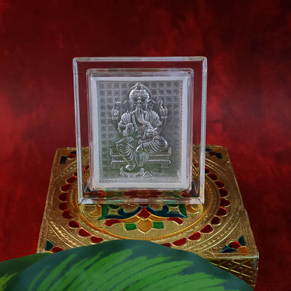 Silver-plated Ganesha idol in an acrylic frame, placed on an ornate gold and multicolored box with a green leaf.