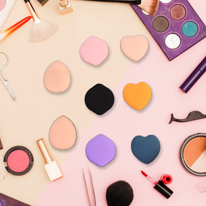 Eight mini makeup sponges in various colors and shapes scattered on a two-tone pink and beige background with makeup.