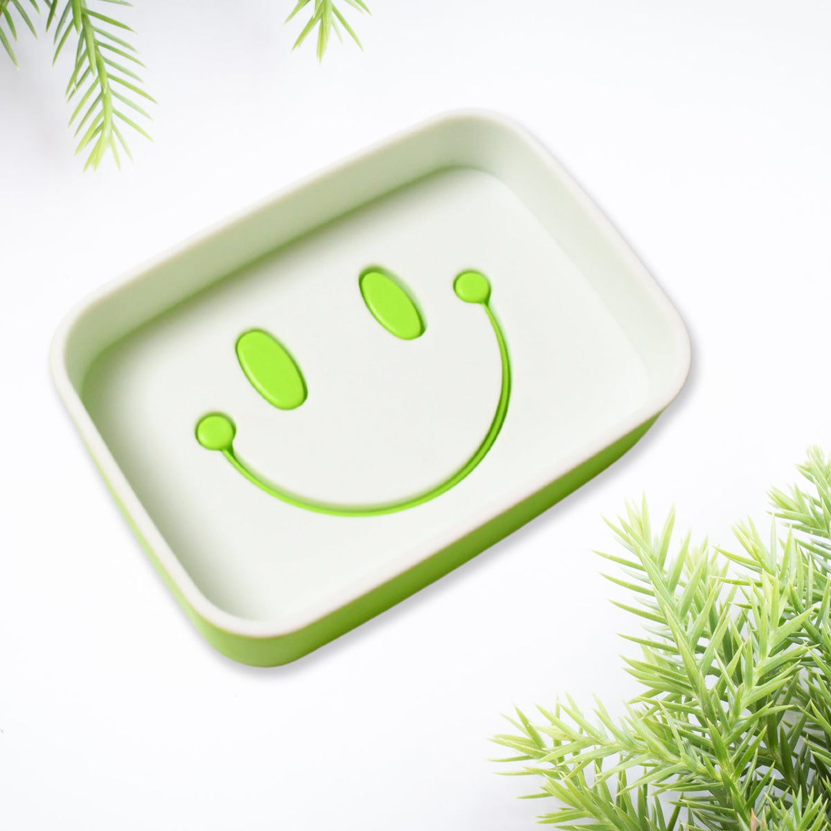 A close-up of a green and white soap dish with a prominent green smiley face draining design.
