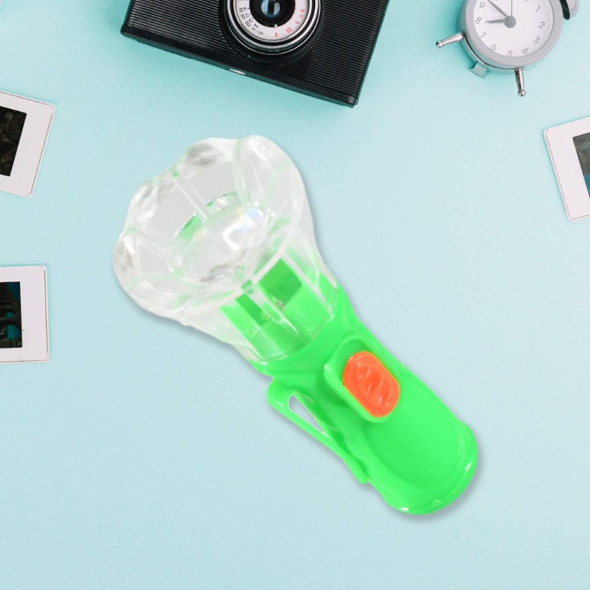 A green and orange plastic flashlight with a clear faceted head is on a light blue surface with a camera and alarm clock.