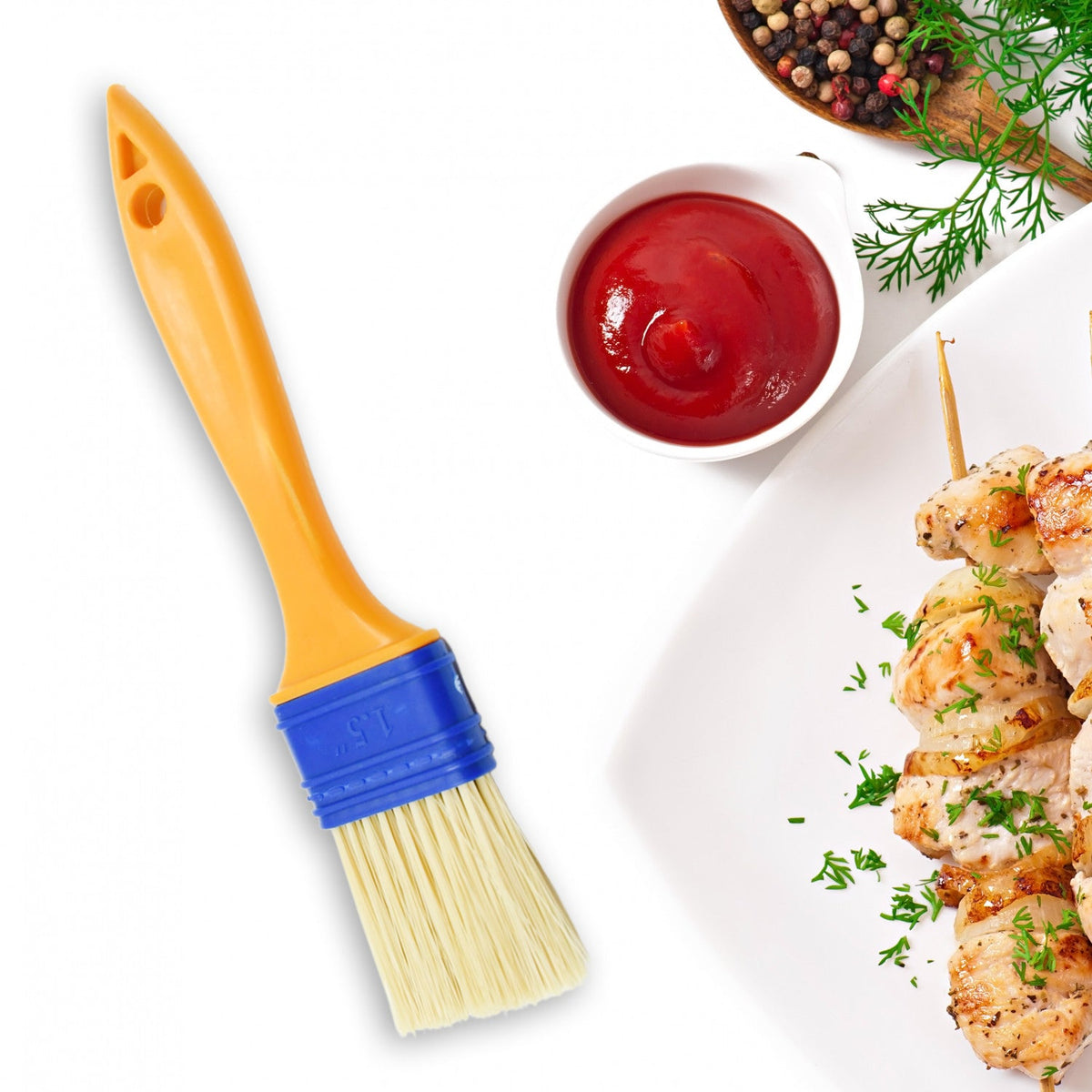 A yellow and blue basting brush is positioned next to a bowl of red sauce and skewers of grilled chicken.