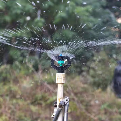 A black and green sprinkler nozzle spraying water in a garden, attached to a white pipe.