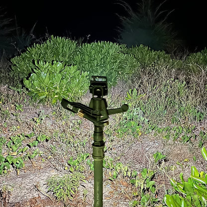 A black mini LED flashlight on a tripod stand, illuminating a dark, leafy outdoor area at night.