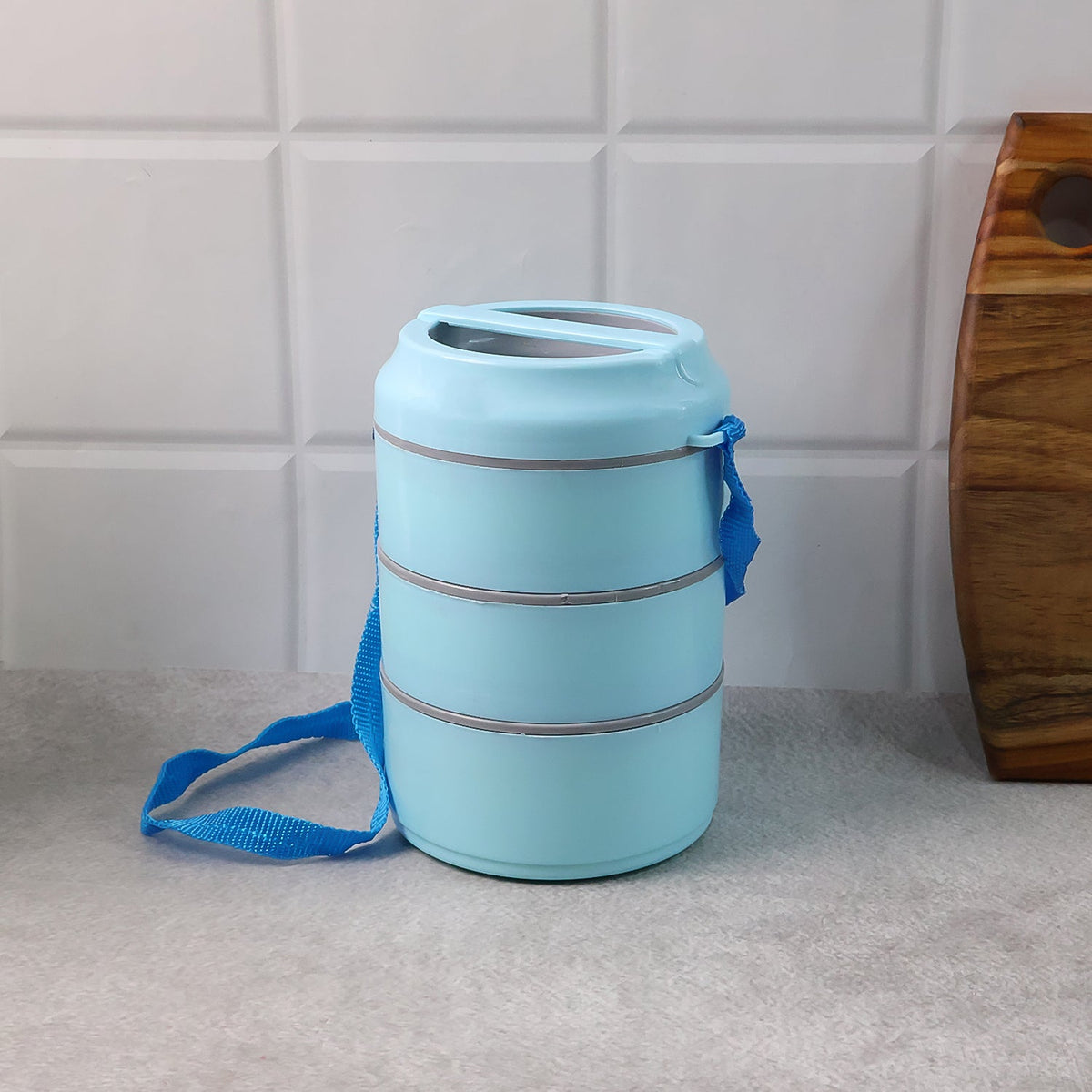 The light blue three-layer lunch box with its blue strap is displayed on a grey countertop against a white tiled wall.