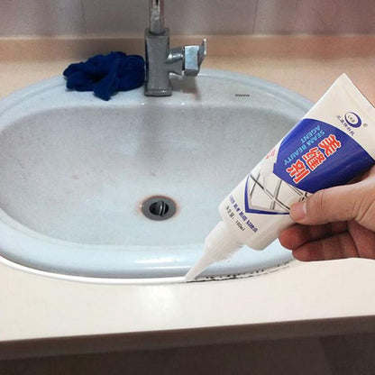 A hand applies white sealant from a tube to the edge of a white bathroom sink, sealing a dark gap.