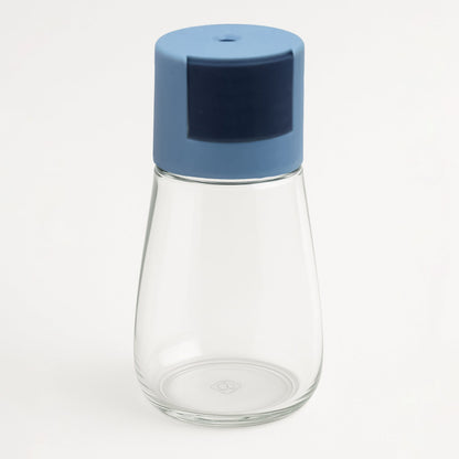 A clear glass spice dispenser with a blue top stands empty on a plain white background.