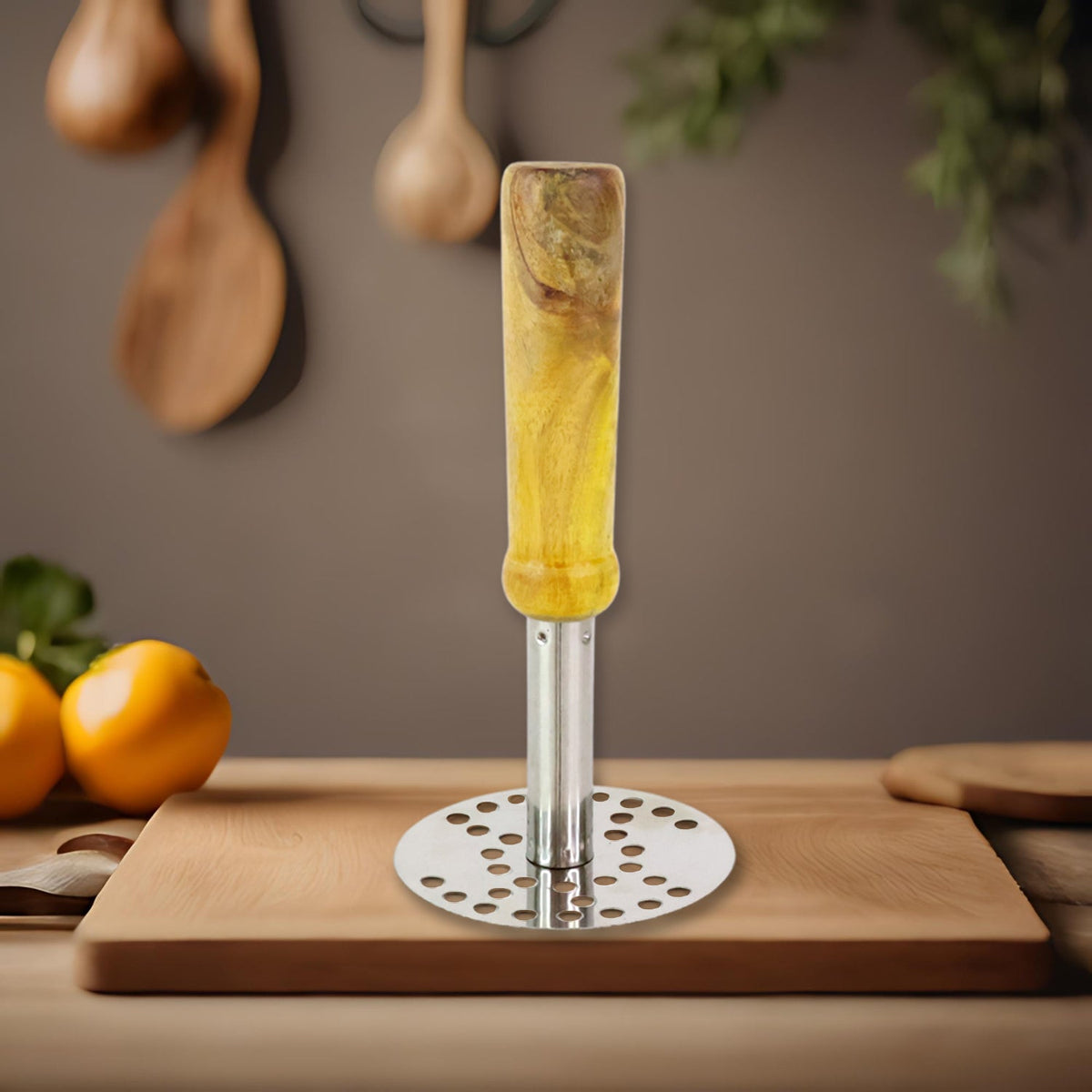 A stainless steel masher with a wooden handle stands on a light wooden cutting board, with lemons and utensils in the
