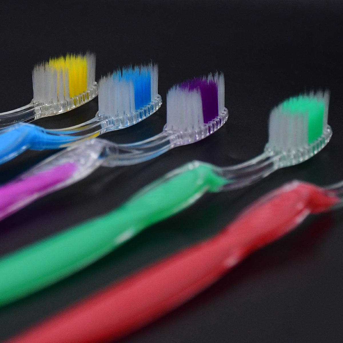 Close-up of the colorful bristles and clear plastic handles of five toothbrushes on a dark surface.