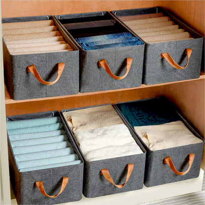 Six grey fabric storage boxes with brown handles, filled with folded clothes, on light wooden shelves.
