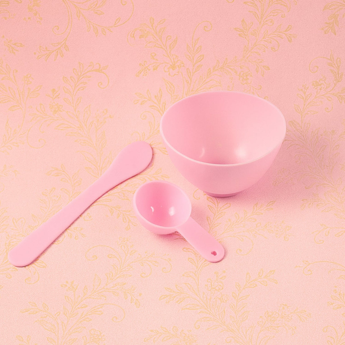 A pink mixing bowl, spatula, and measuring spoon are arranged on a pink background with a subtle floral pattern.