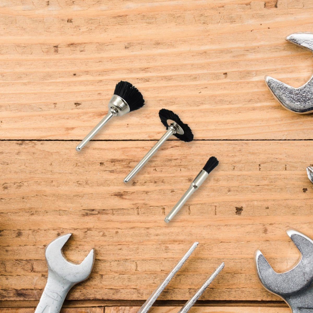 Three black nylon brushes with silver shafts are arranged on a light wooden surface surrounded by silver wrenches.