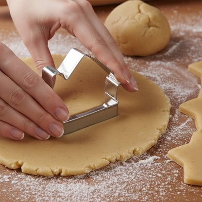 Hands using a house-shaped cookie cutter on rolled-out dough, with a ball of dough and star-shaped cutouts visible.
