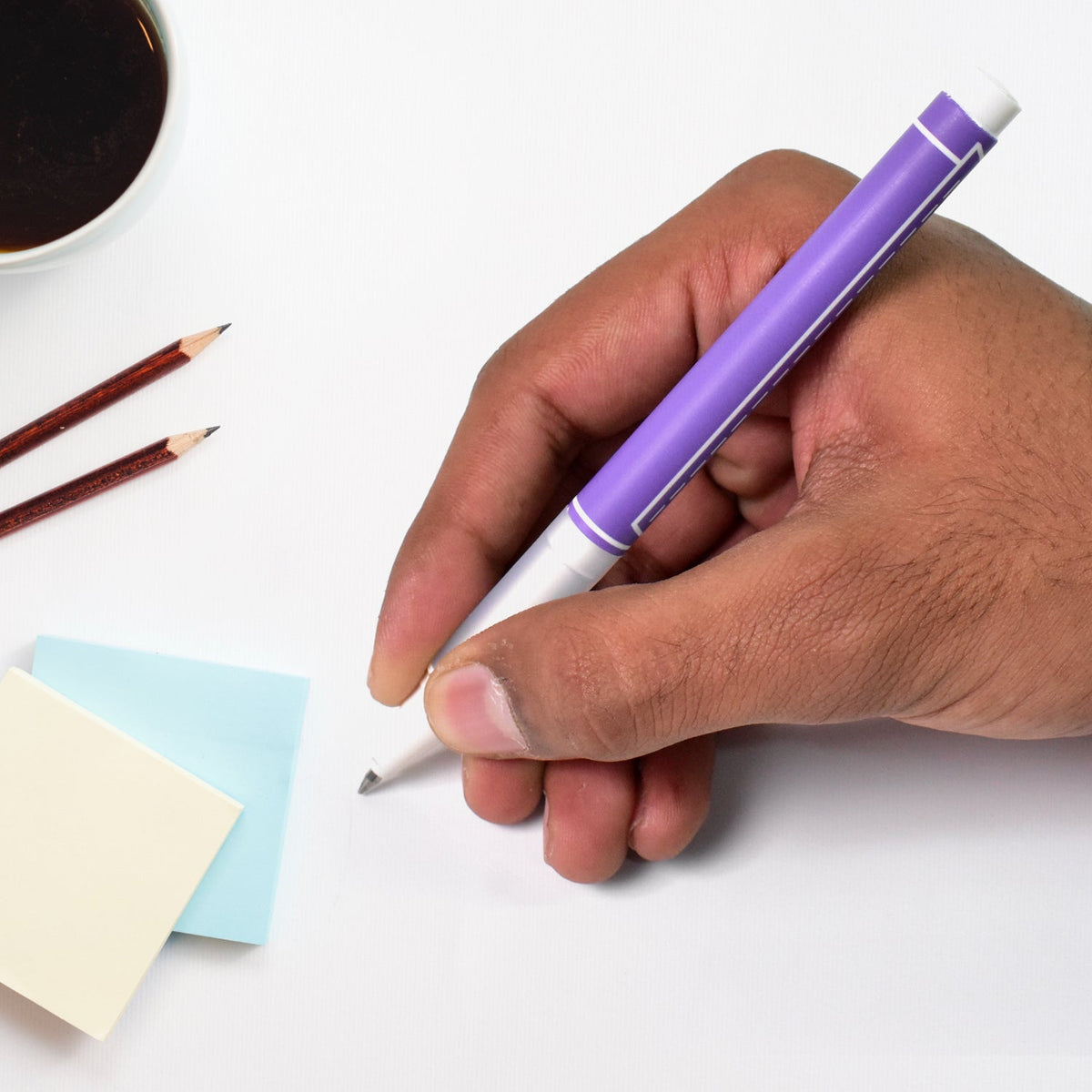 A hand holds a purple gel pen, writing on a white surface next to sticky notes and pencils.